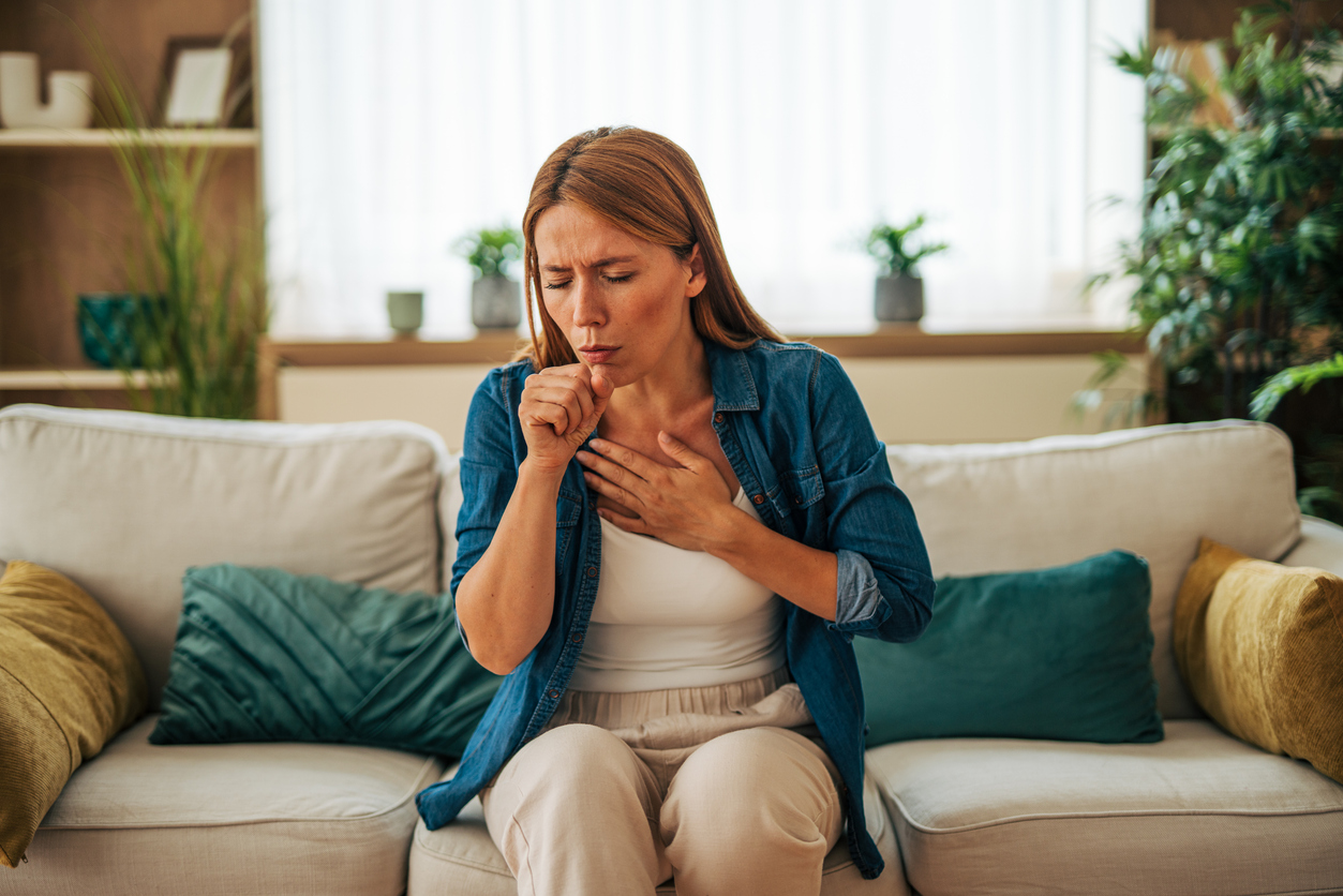 Sick woman coughing and holding chest while sitting on sofa at home